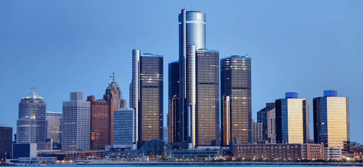 Photograph of Detroit skyline at dusk from across the Detroit River featuring The Renaissance Center and the surrounding shorter rectangular skyscrapers and buildings are illuminated by setting sunlight.
