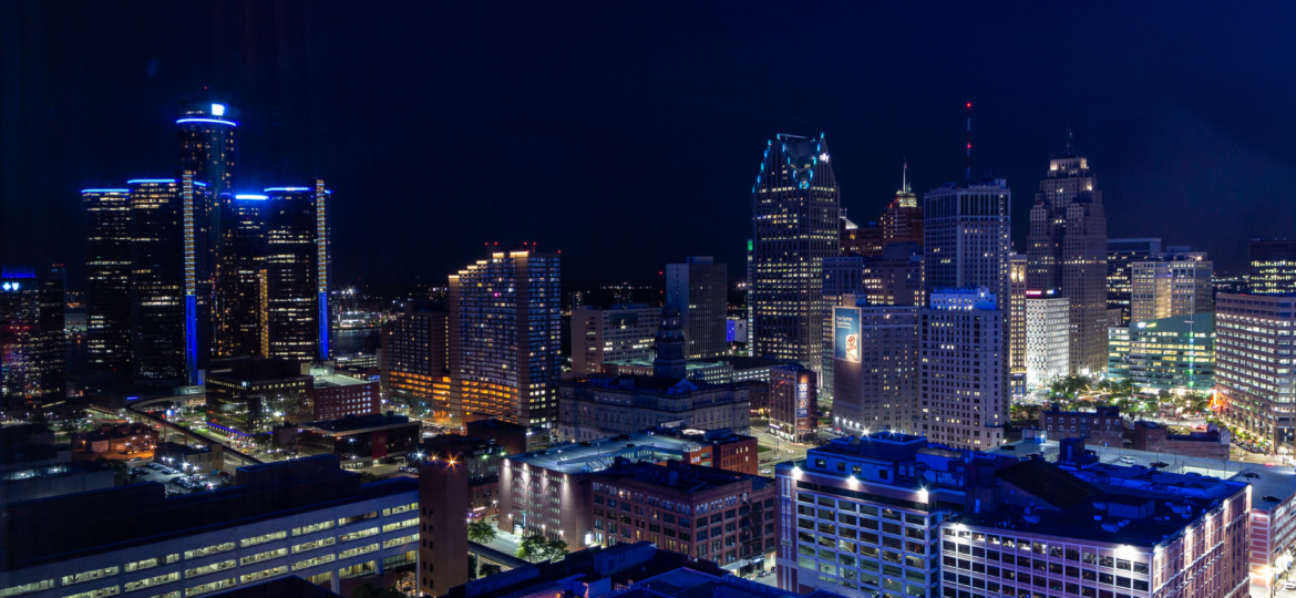 Downtown Detroit skyline at night showcasing city lights and illuminated high-rise buildings and office towers against a dark sky.